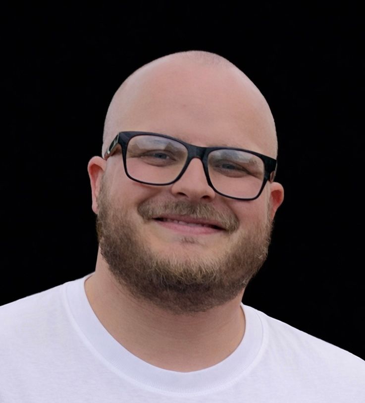 Man with glasses smiling, wearing a white t-shirt, against a black background.