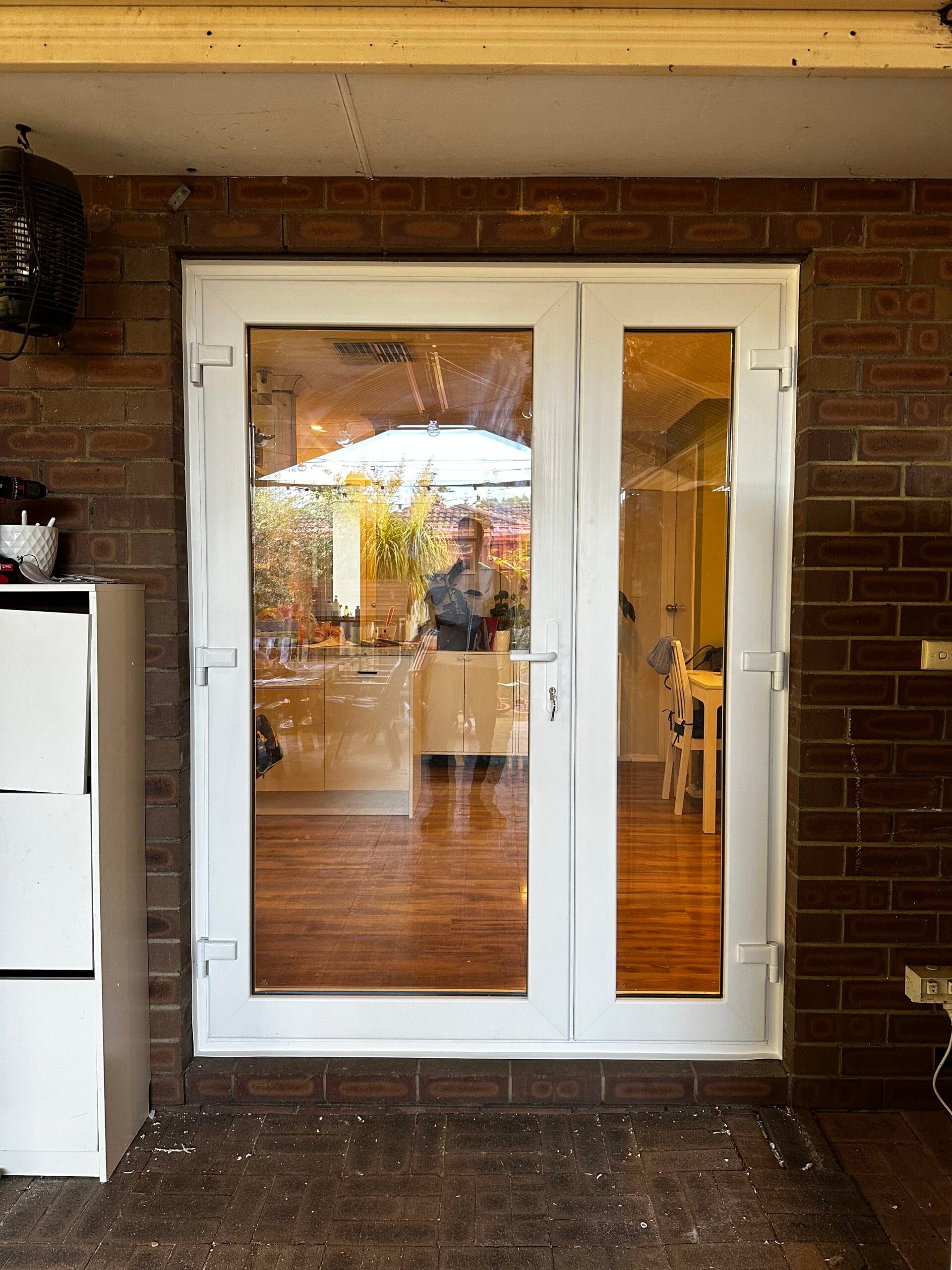 White framed French doors with glass panes reflecting a kitchen, set in a brick wall.