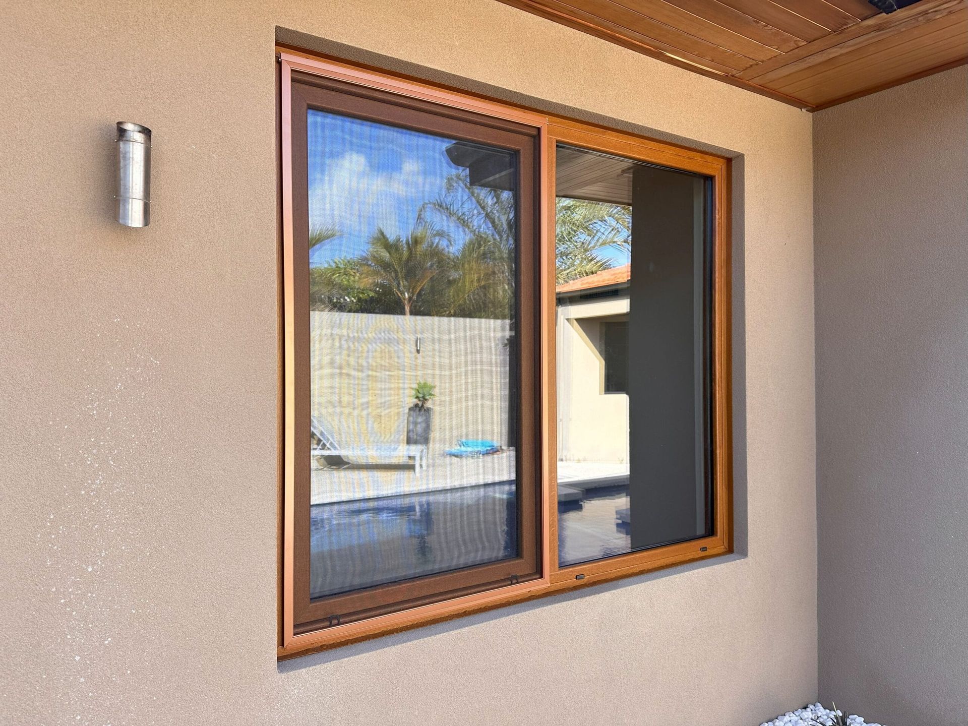 Brown-framed window with screen on stucco wall, reflecting a pool, sky, and building. A silver light fixture is on the wall.