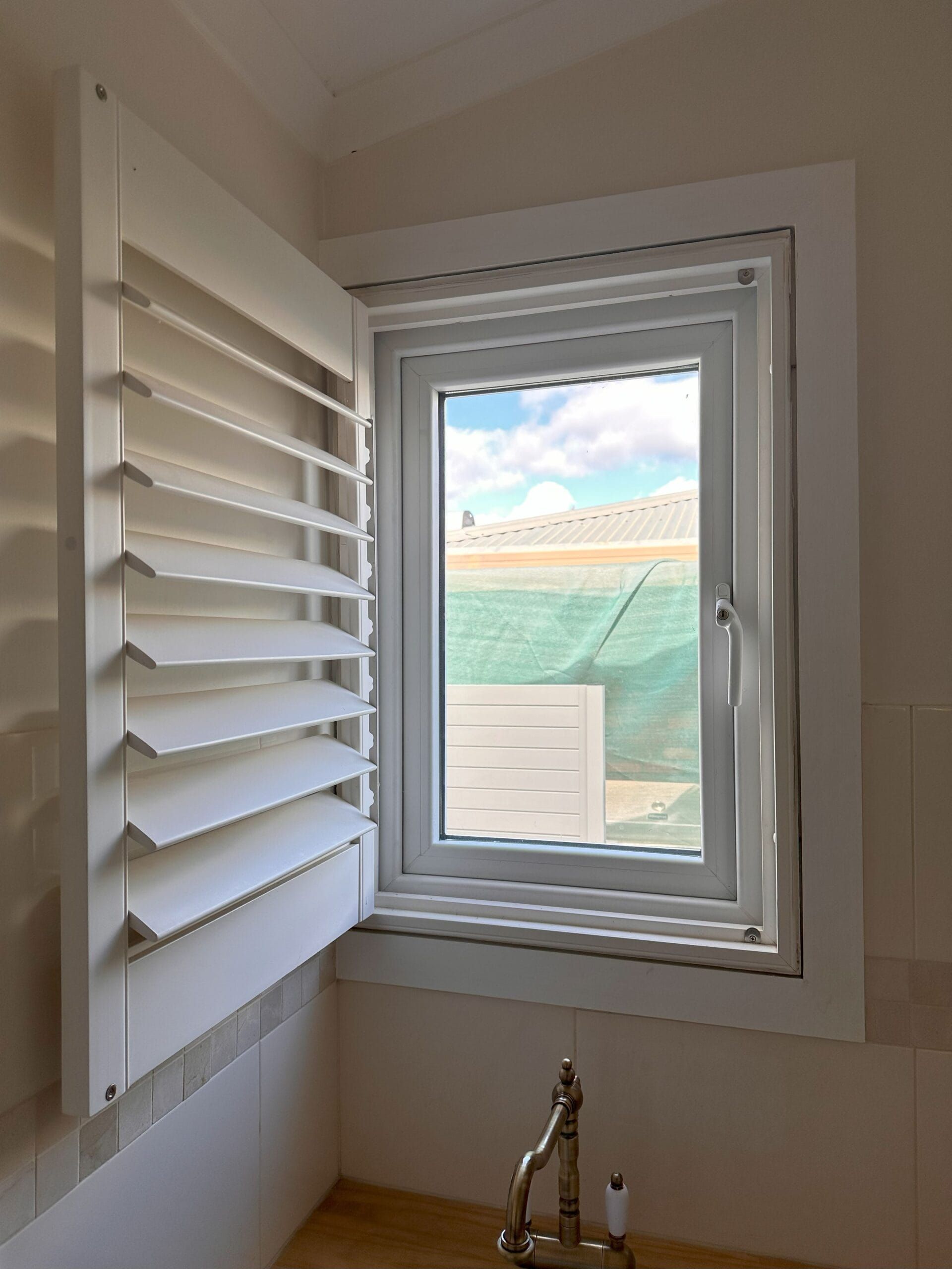 White window with open shutter, revealing a view of rooftops and a green mesh fence.