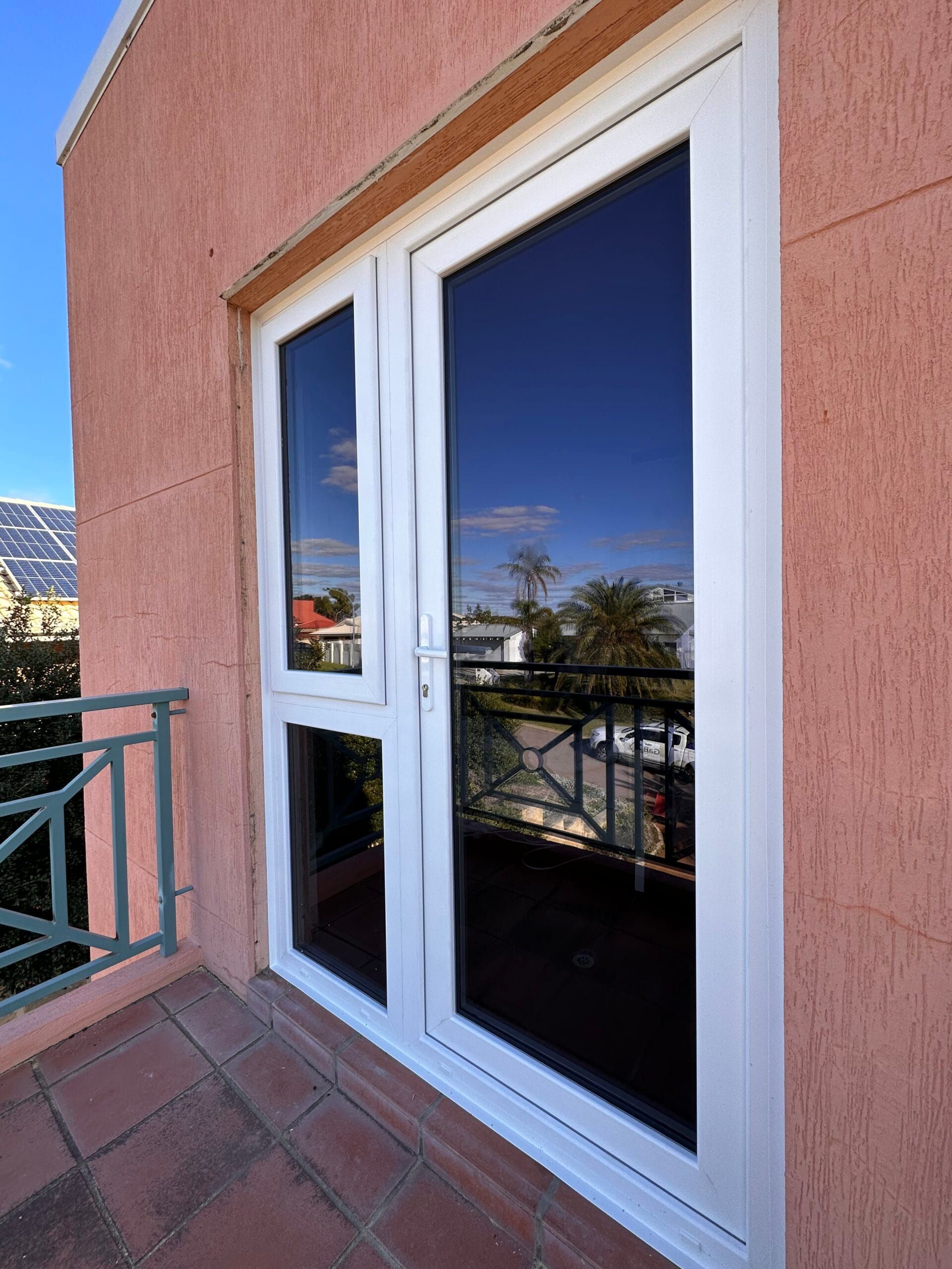 White-framed double doors with reflective windows on a pink stucco wall, leading to a balcony with green railing.