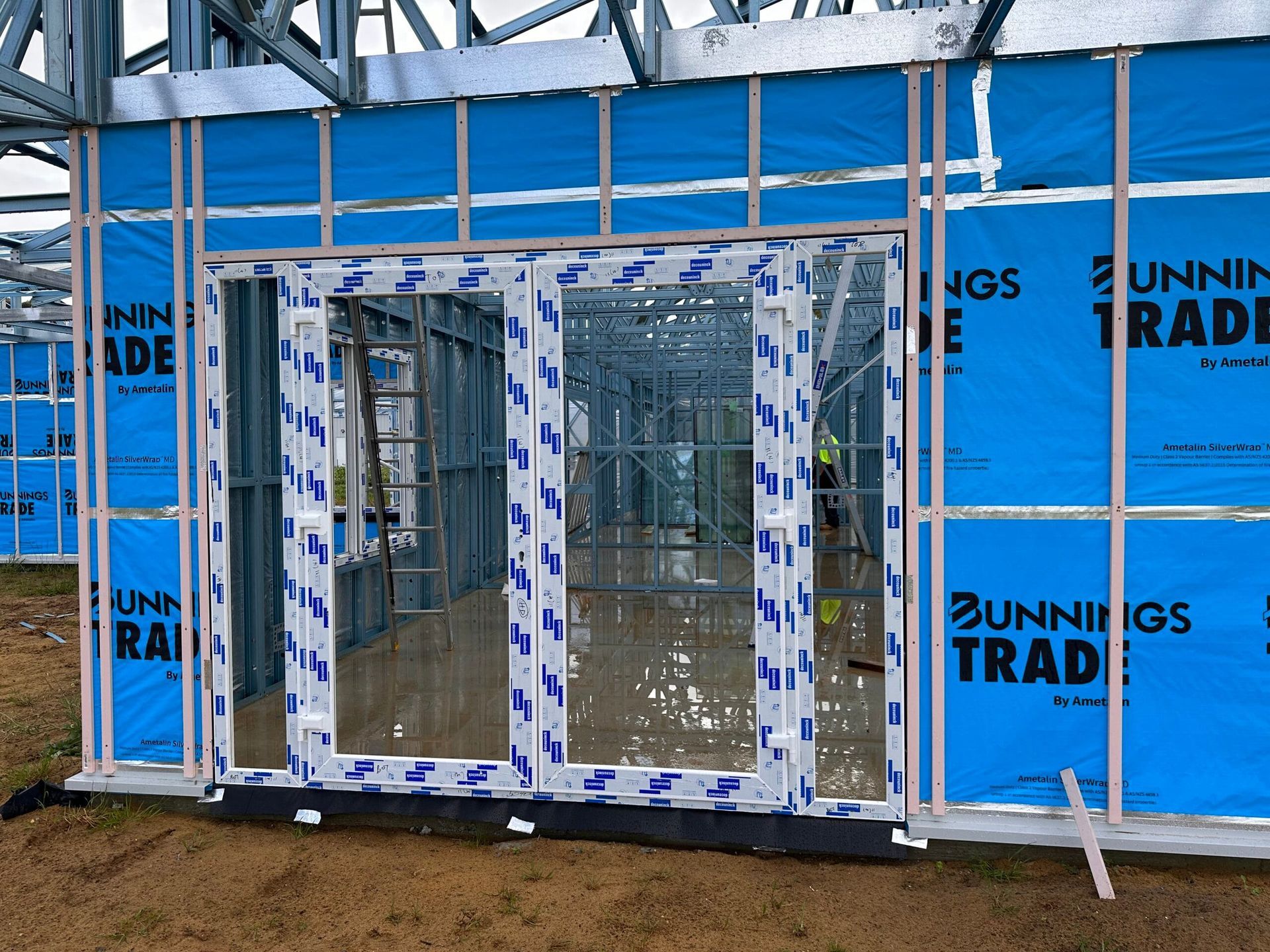 Construction site: metal frame building with glass doors, blue insulation, and Bunnings Trade branding.