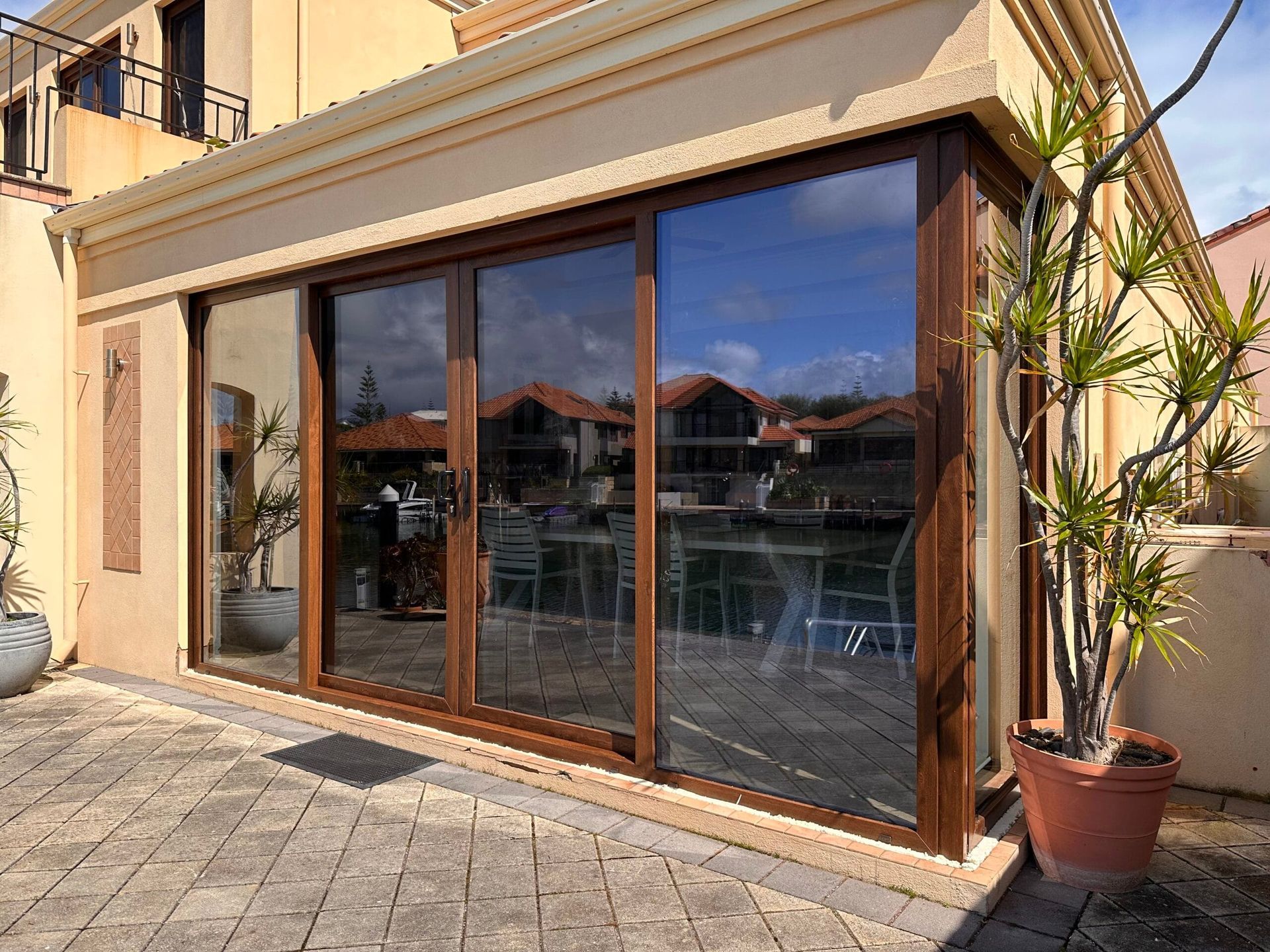 Brown sliding glass doors on a tan building, overlooking water and homes. A potted plant stands to the right.