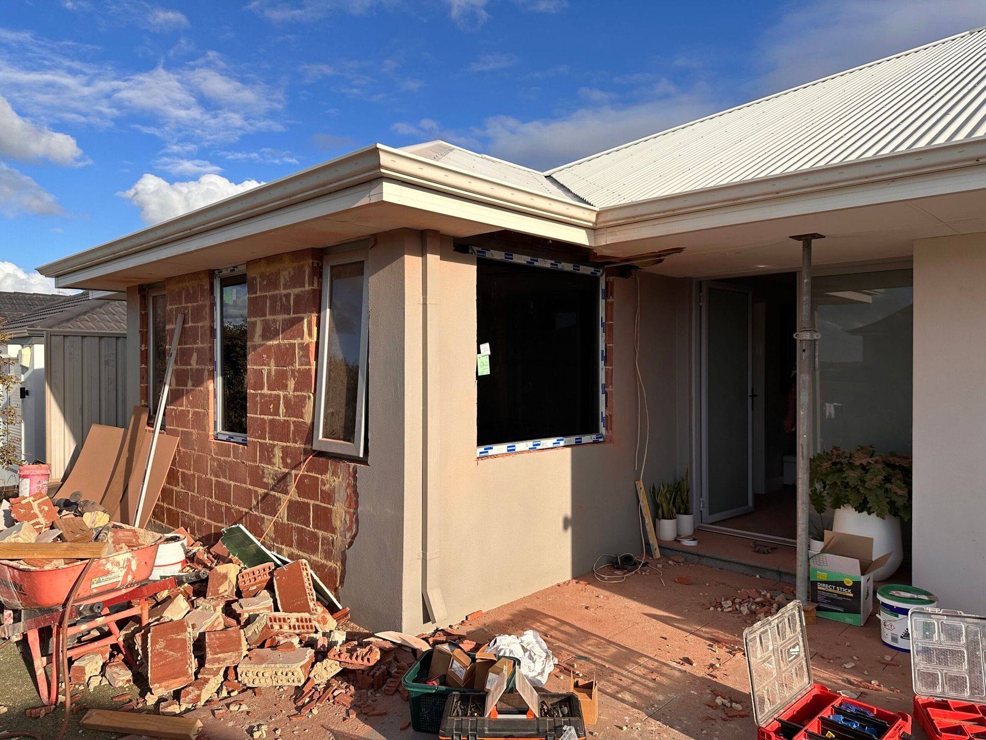 House exterior under construction, exposed brick, stucco, with windows and support beams. Construction tools and debris visible.