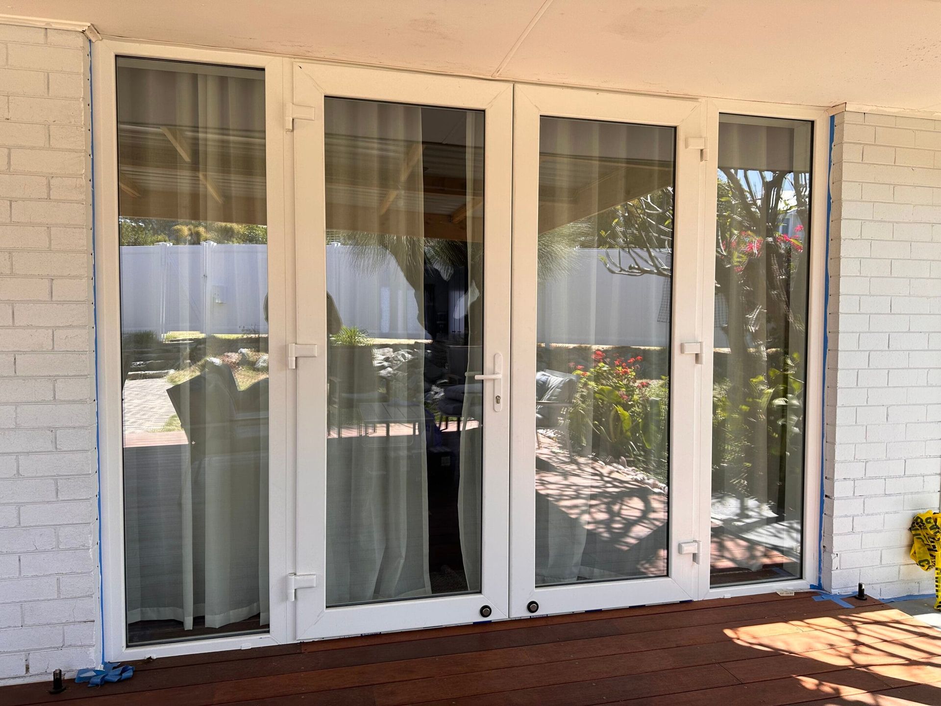 White framed French doors with sheer curtains, reflecting an outdoor view, on a brick wall.