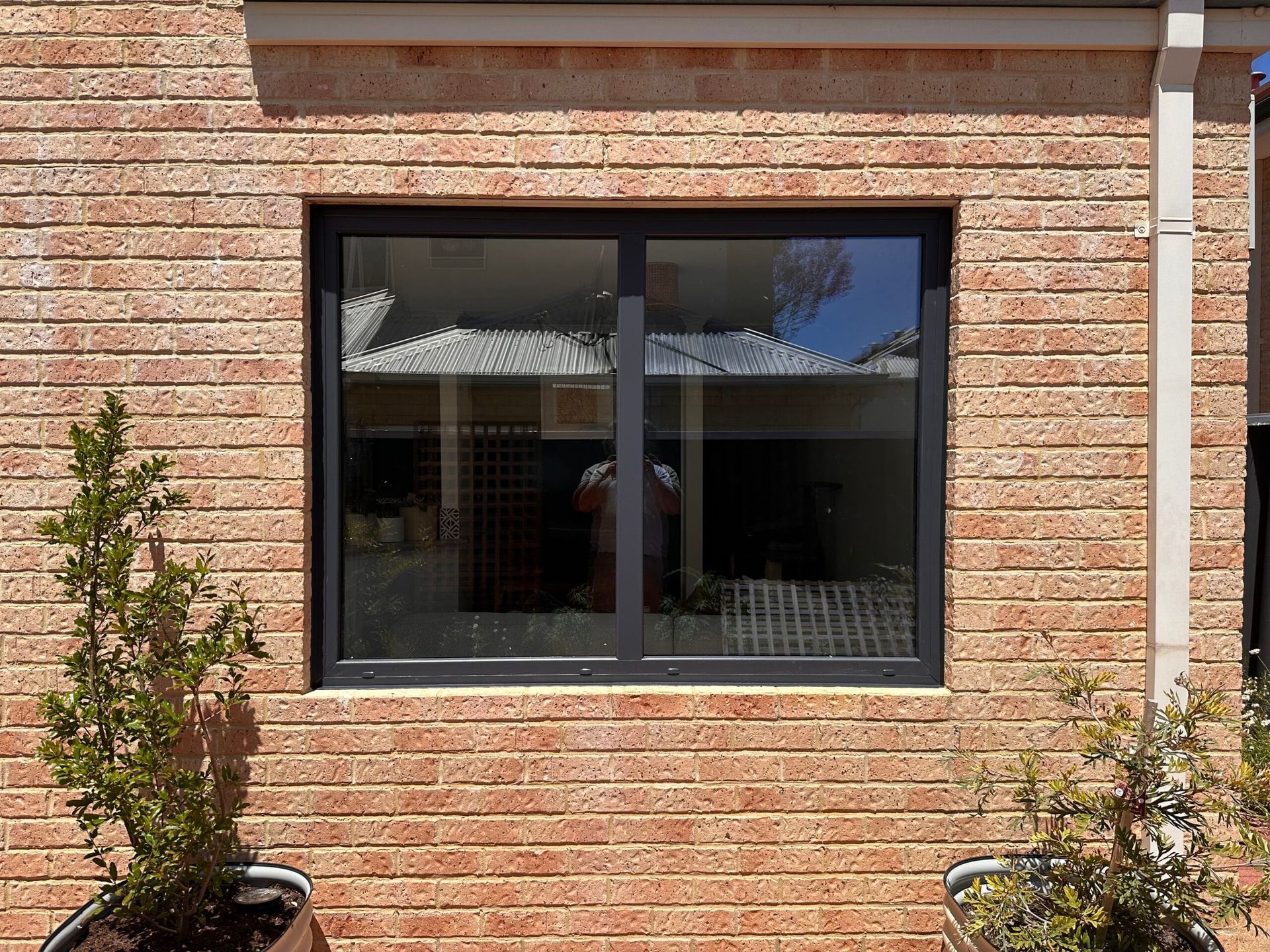 Brick wall with a dark-framed window reflecting a building and a downspout.