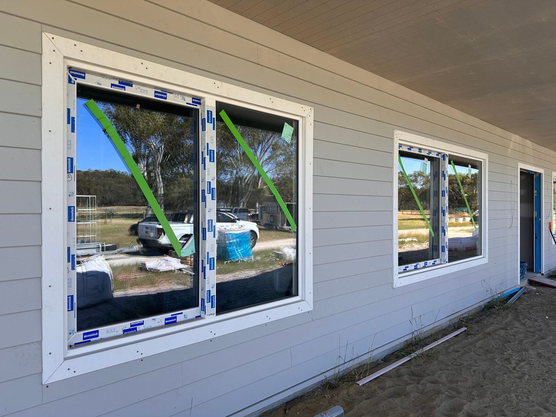 Three white-framed windows on the exterior of a light gray house under construction; reflective glass; green tape.