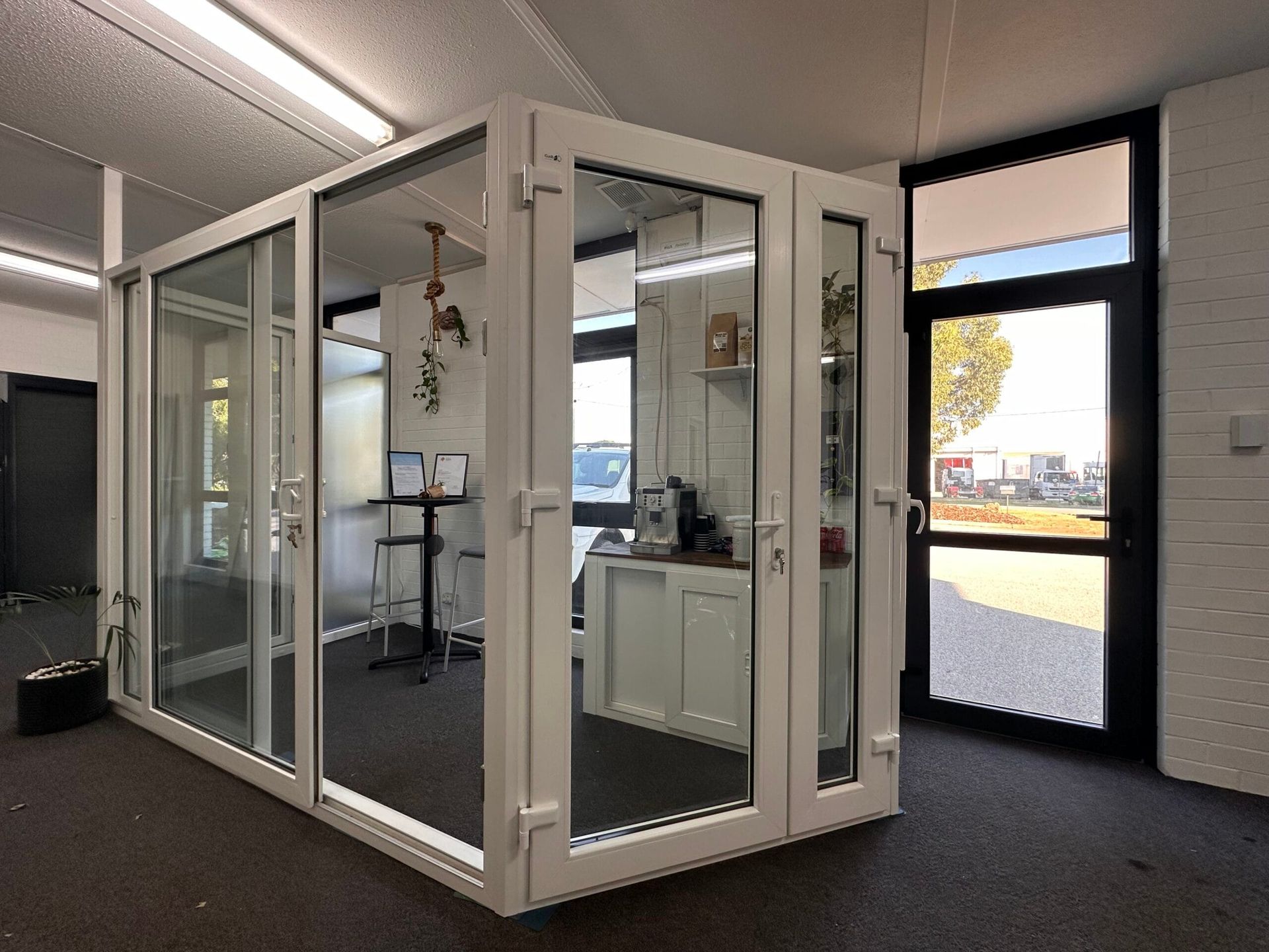 White-framed glass enclosure with door, inside a room; black door and exterior visible.