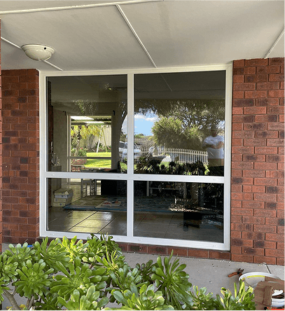 White-framed window in brick building, reflecting outdoor scenery including plants and a lawn.