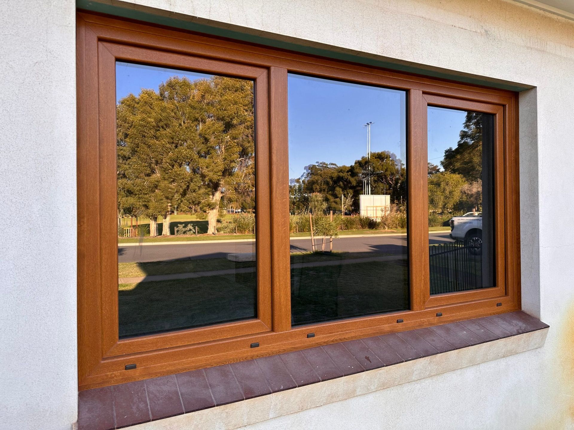 Brown-framed triple-pane window reflecting a street, trees, and blue sky. Set in a white stucco wall.