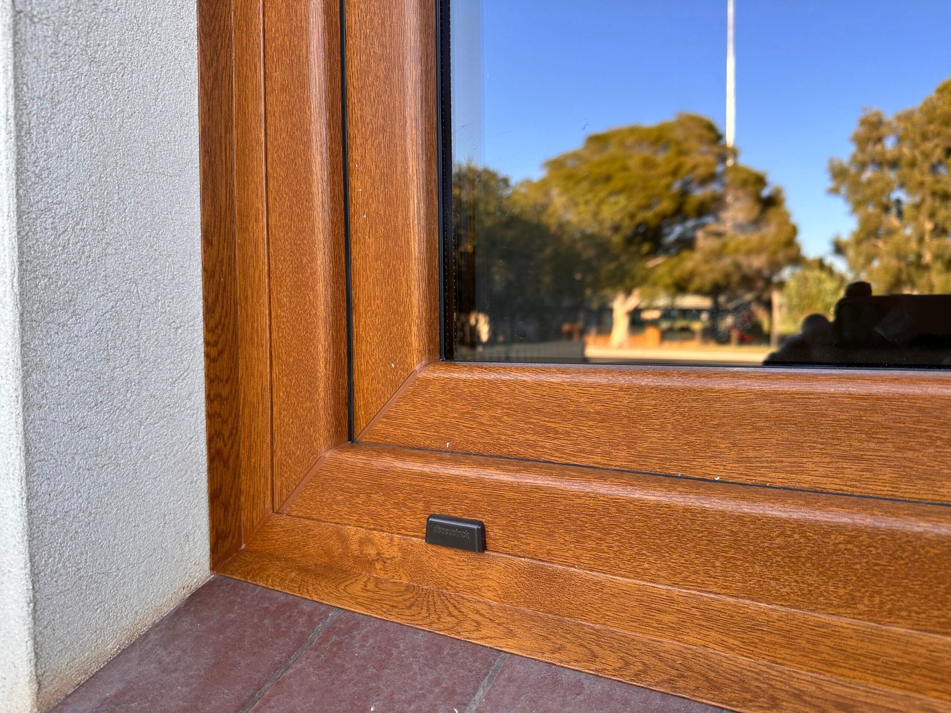 Brown window frame with a textured finish, set in a corner with a grey wall and visible outdoor trees.