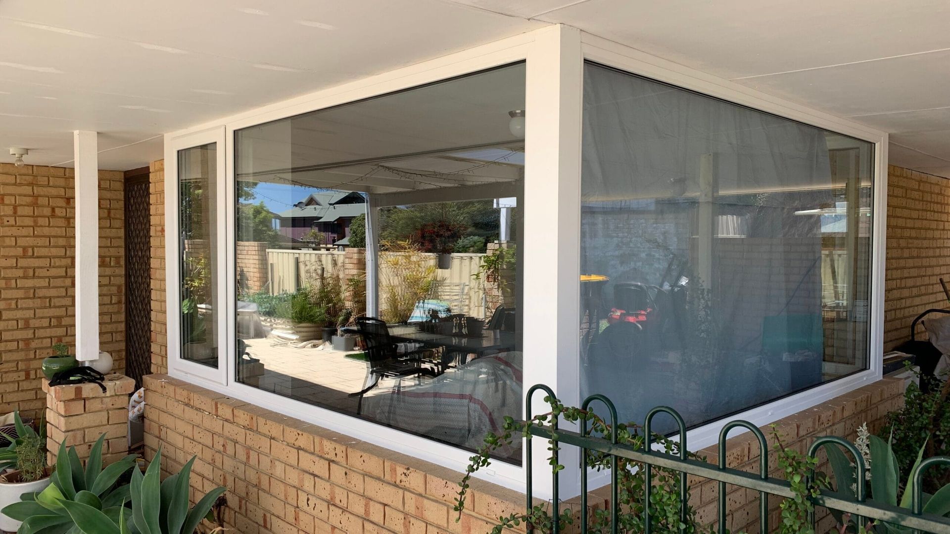 Glass-enclosed porch with white trim and large windows, reflecting outdoor scene and brick base.