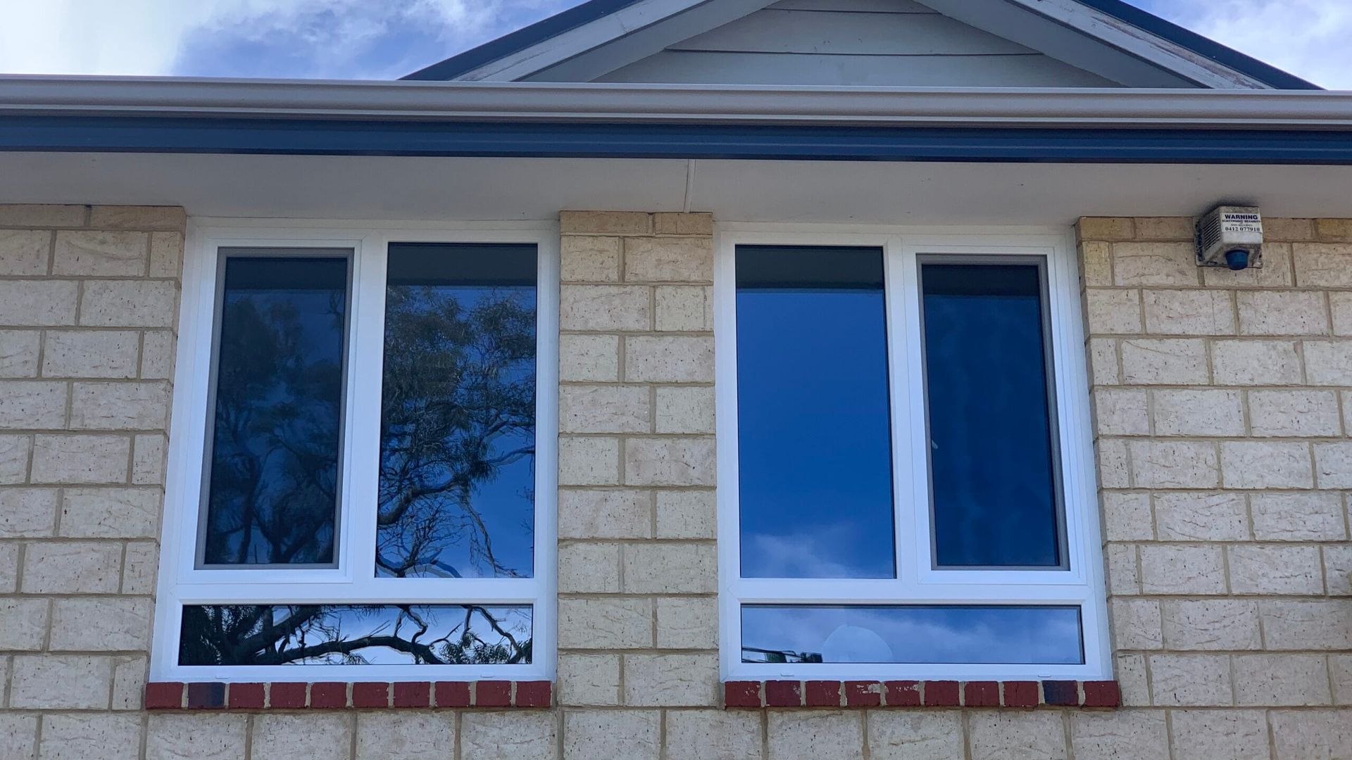 White-framed windows on a brick house reflect the sky and trees. Blue gutter and security camera above.