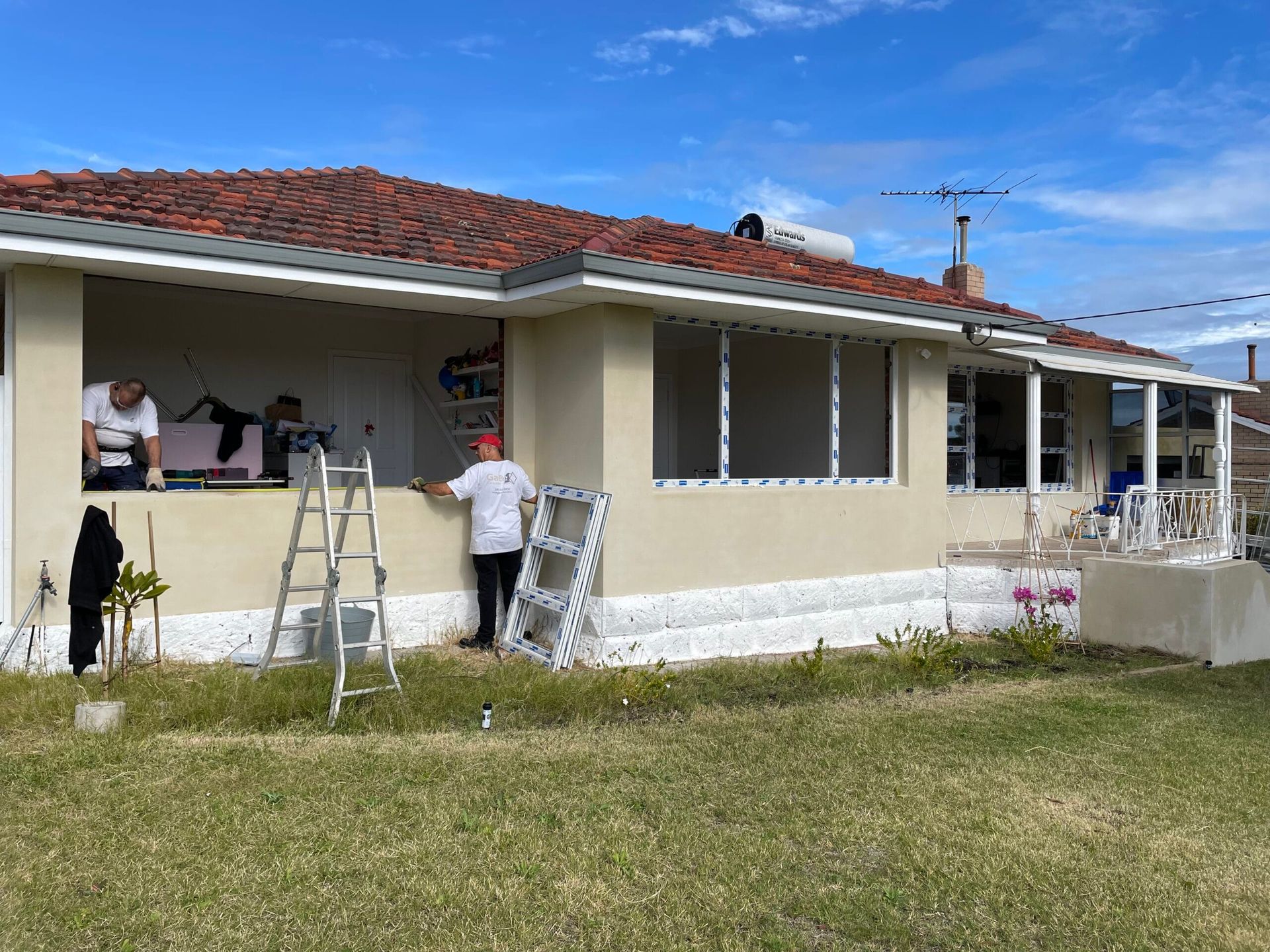 People installing windows on a light-colored, one-story house with a terracotta tile roof, blue sky, and green grass.