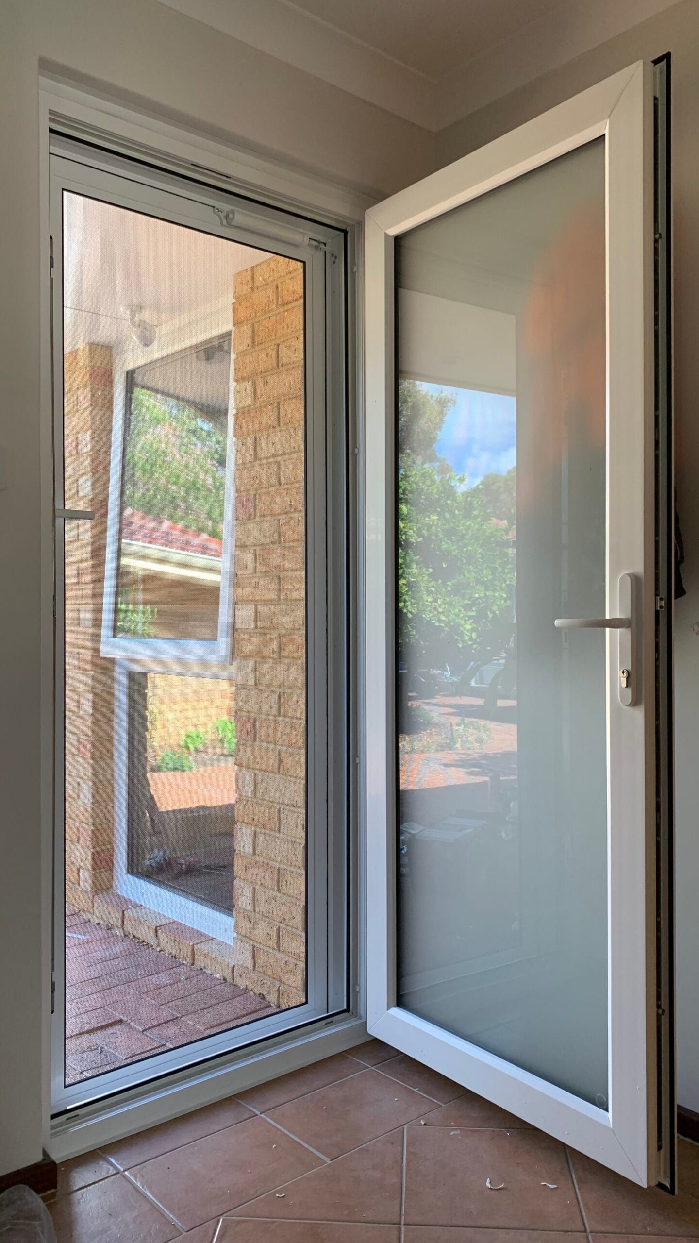 White door ajar, with frosted glass, revealing outdoor view. Adjacent open window, brick wall.