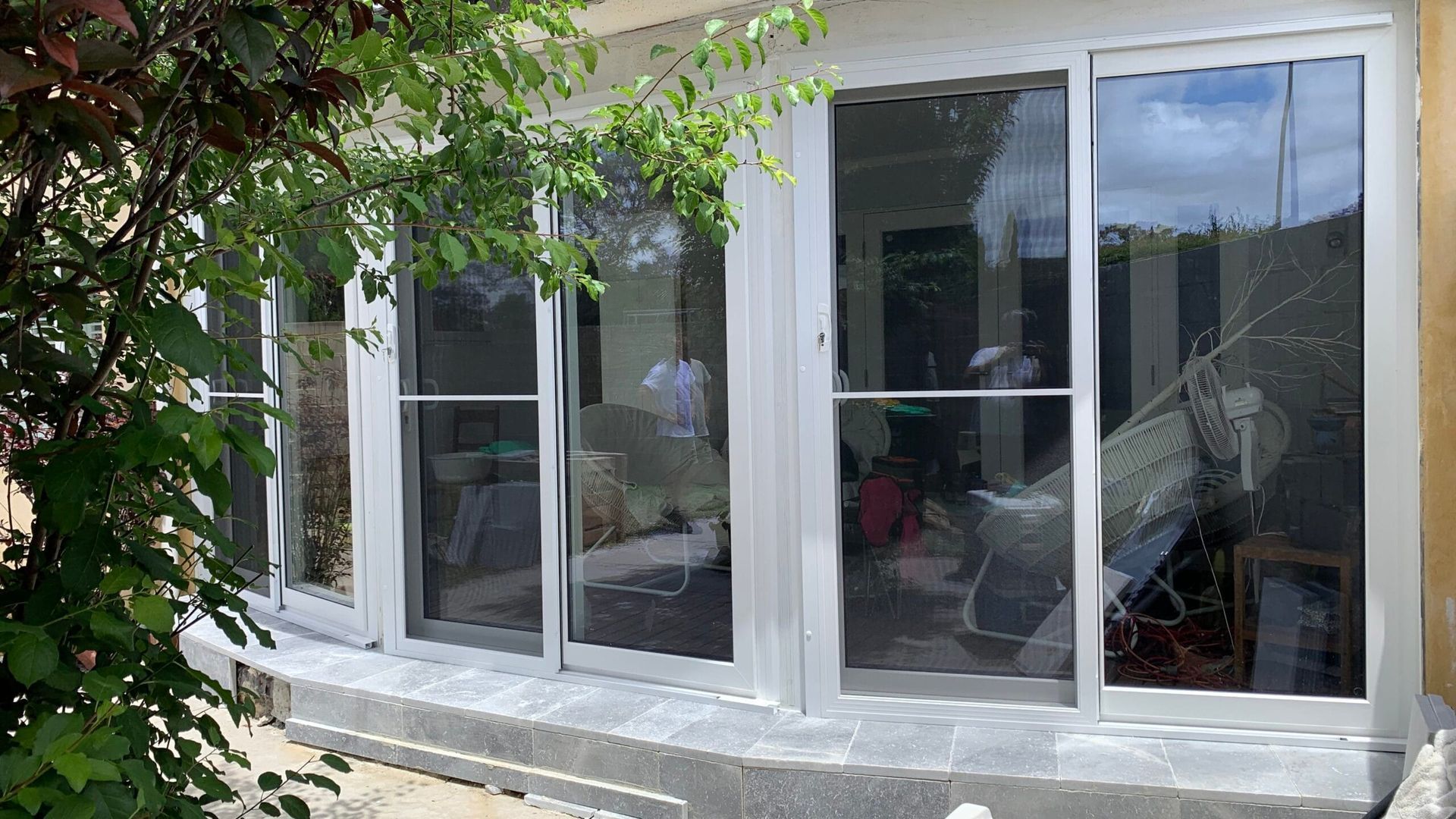 Exterior view of house with large windows, silver frames, and stone ledge. Green foliage visible on left.
