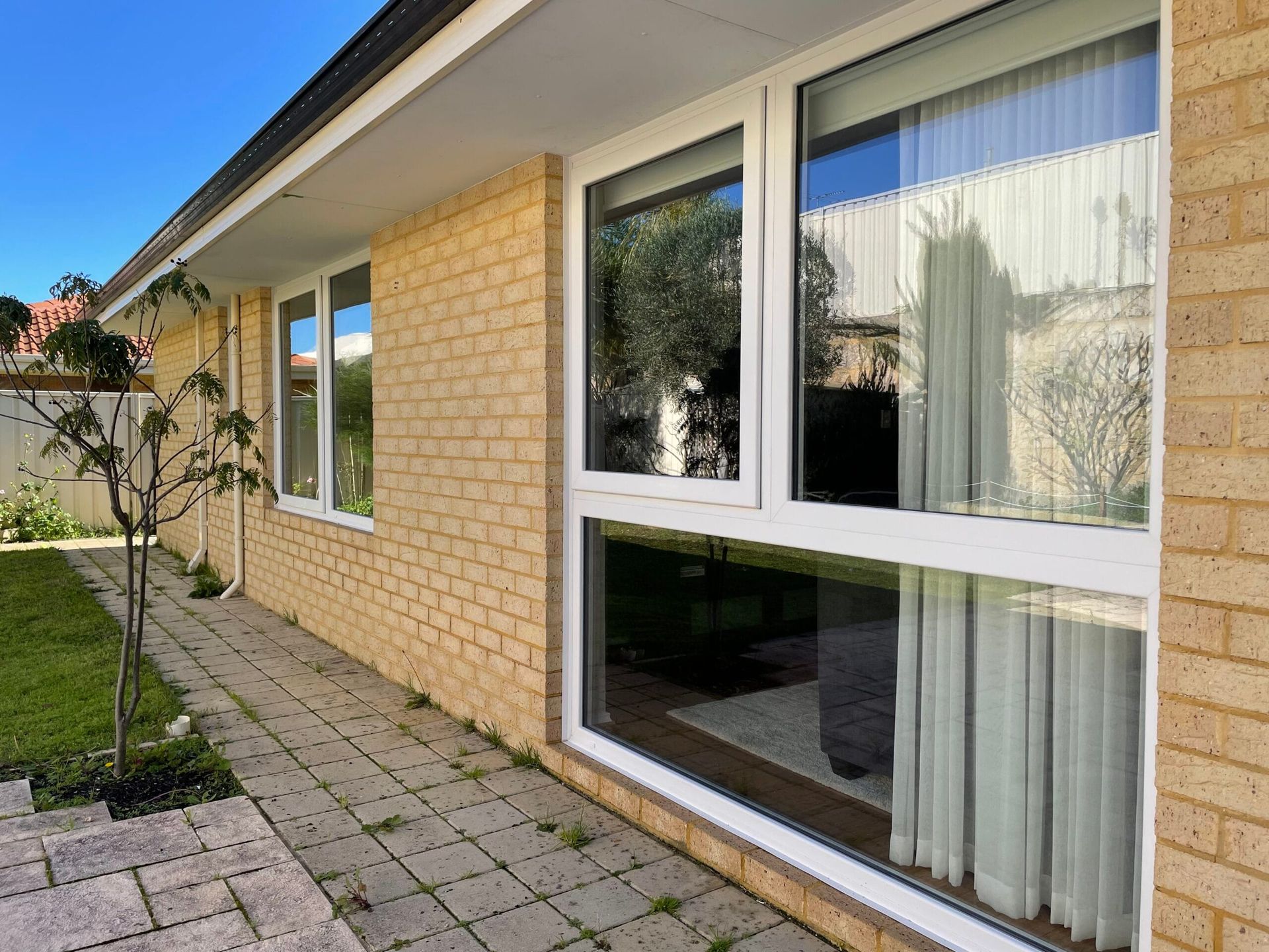 Exterior of a light brick house with white-framed windows reflecting the surroundings, including a courtyard and plants.