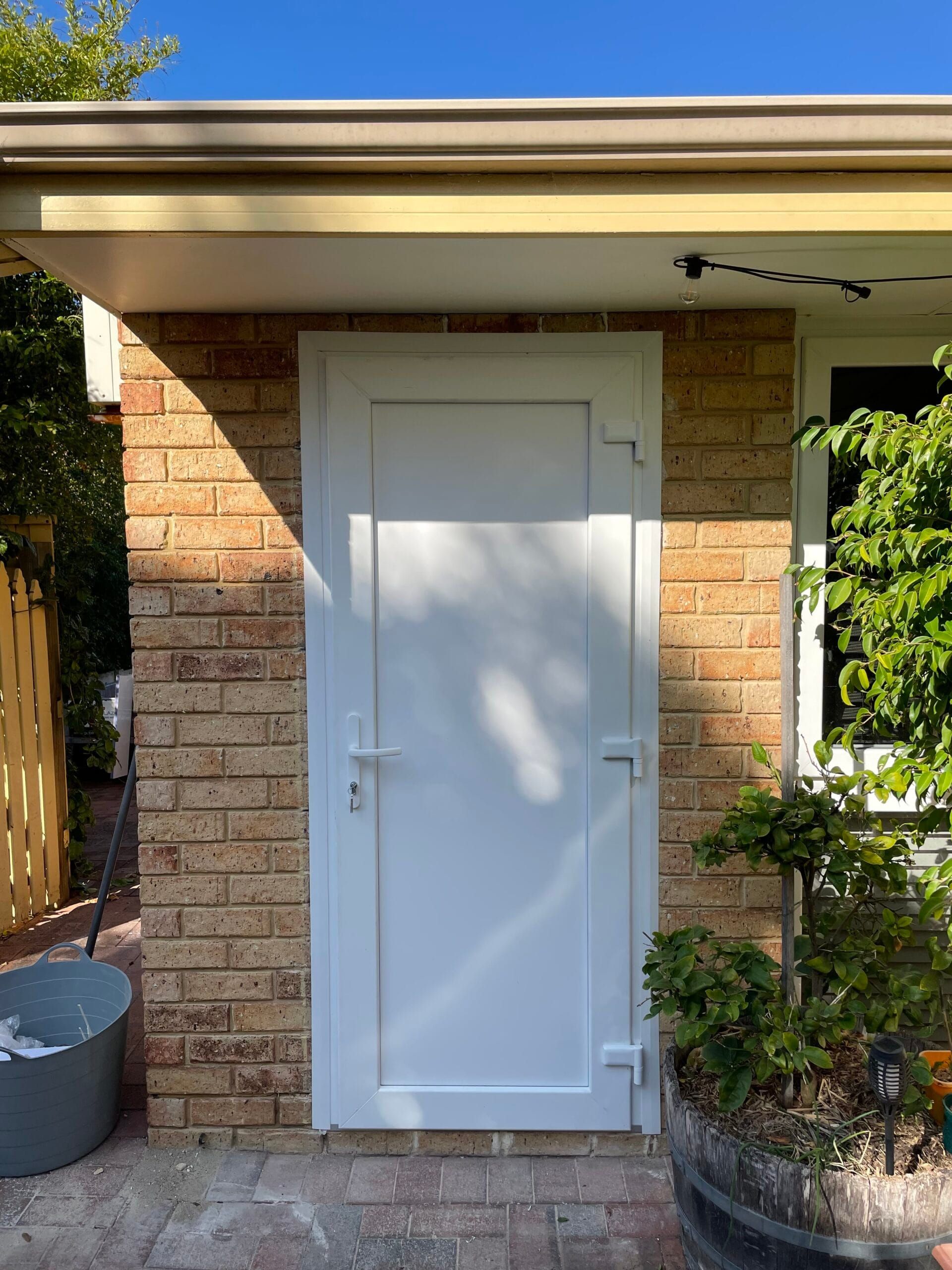 White door set in a brick wall, under a tan roof overhang; sunlight.