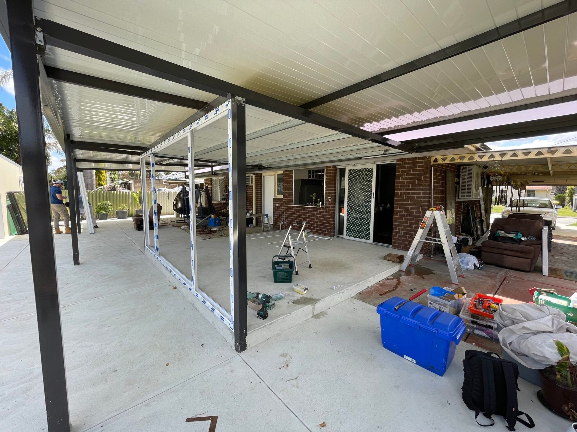 Construction of a patio with a metal frame and partially enclosed glass walls. Tools and materials are scattered around.