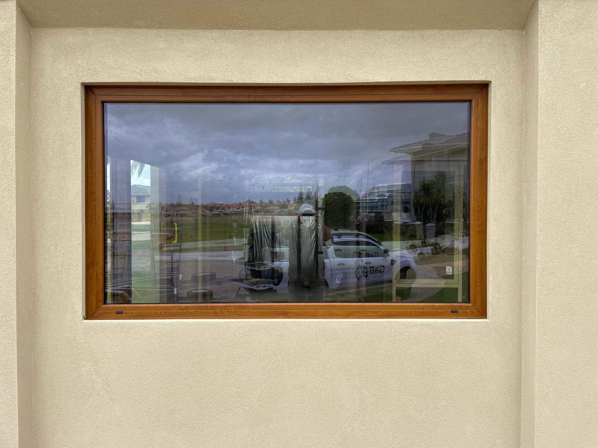 Brown-framed window reflecting an overcast sky, buildings, and vehicles, set in a beige wall.
