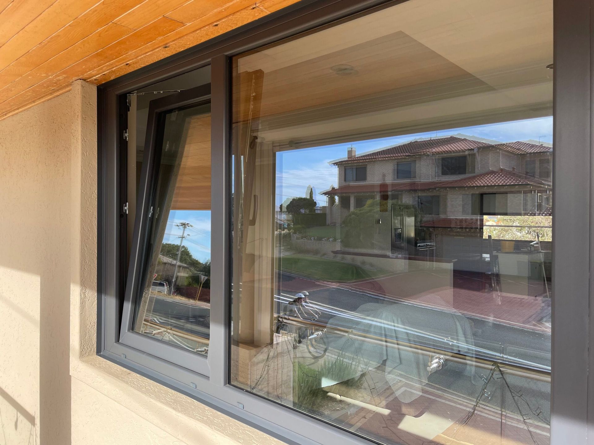 Open gray tilt-and-turn window reflecting house and sunny sky. Beige wall.