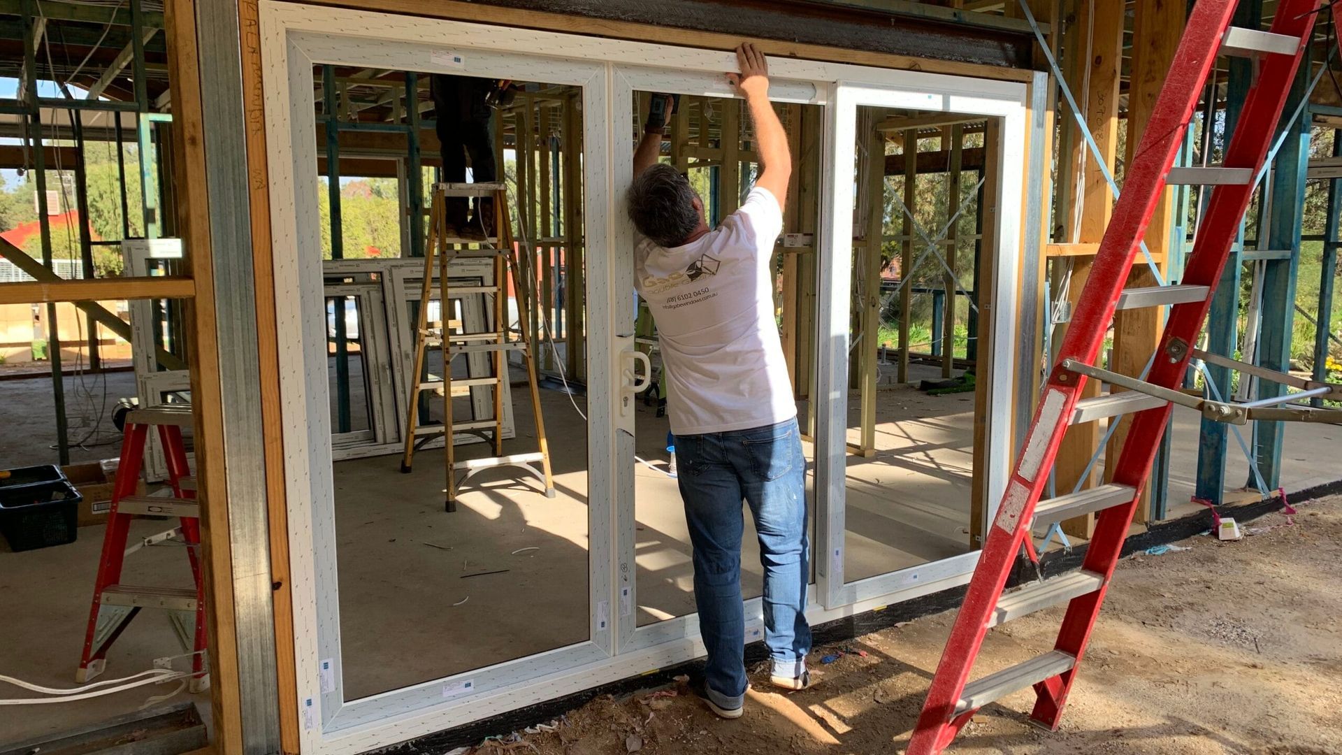 Man installing a sliding glass door in a house under construction; a ladder and a second person on another ladder are visible.