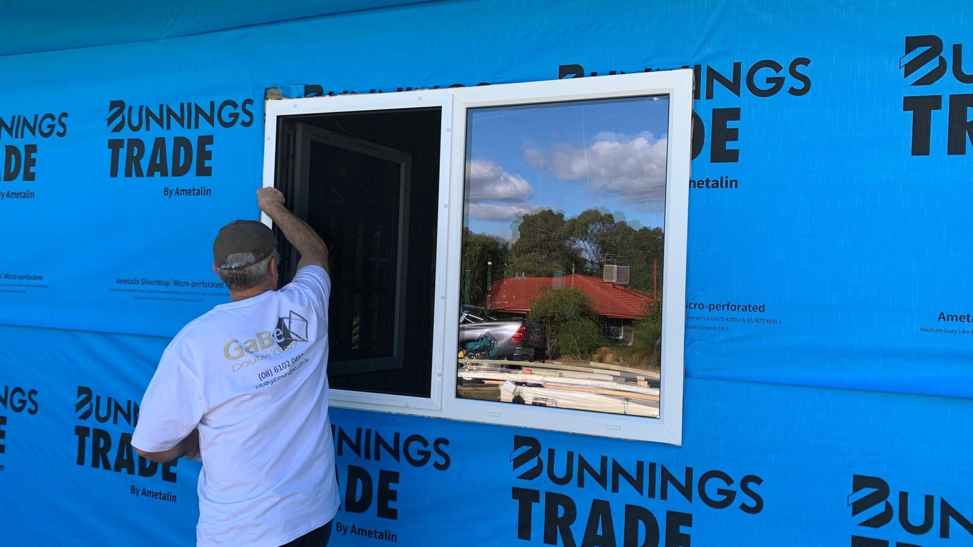 A person installing a white window in a blue-wrapped building, the window reflects a sky, trees, and a red roof.