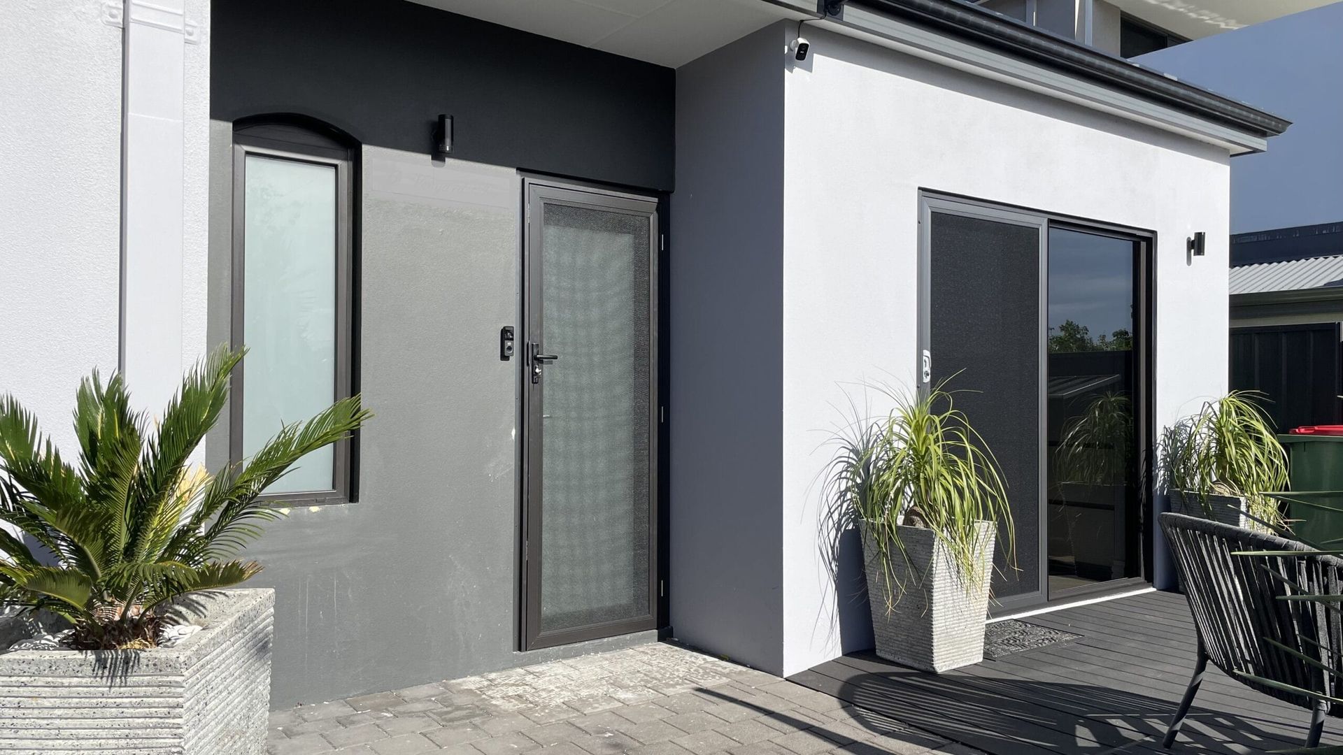 Exterior of a modern home with a door and sliding glass door. Gray and white walls, patio with plants.