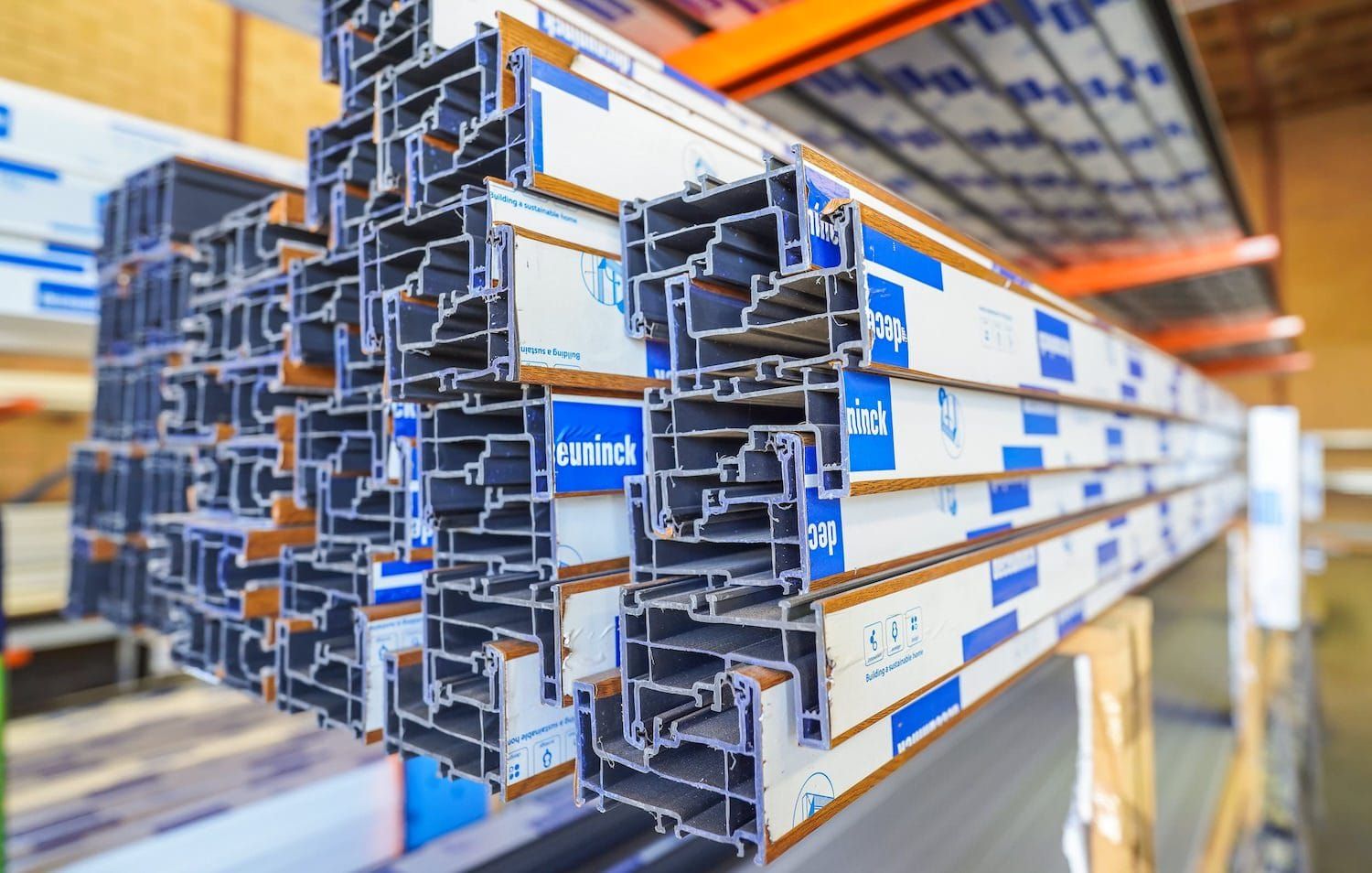 Stacked aluminum window frame profiles in a warehouse, some with blue and white protective wrapping.
