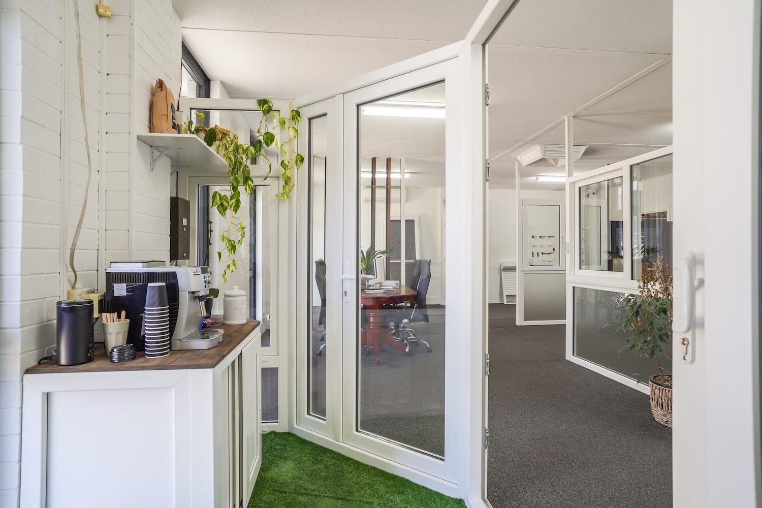 Office interior with white walls, glass doors, and a small green space; view of another office space.
