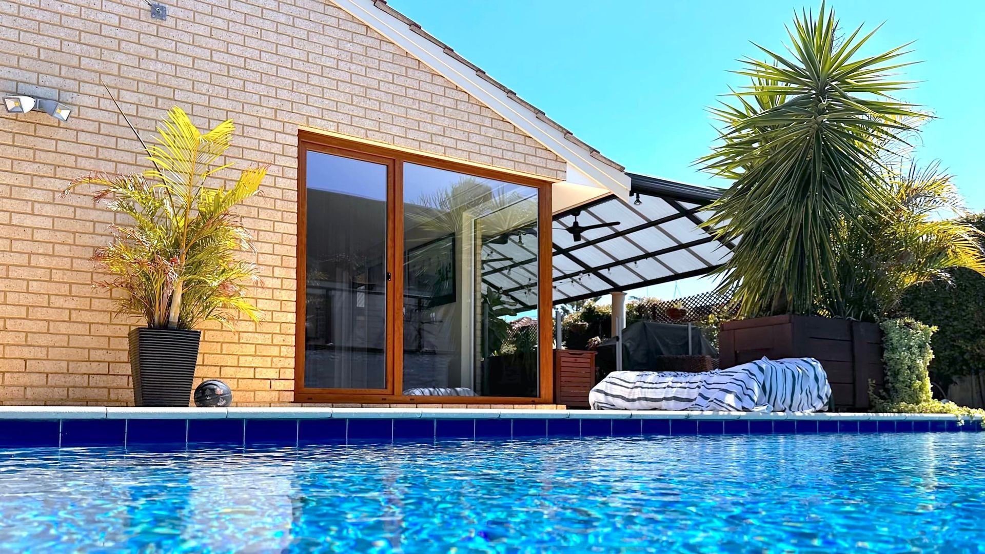 Swimming pool in front of a brick house with large glass doors. Patio and trees in background. Bright blue water.