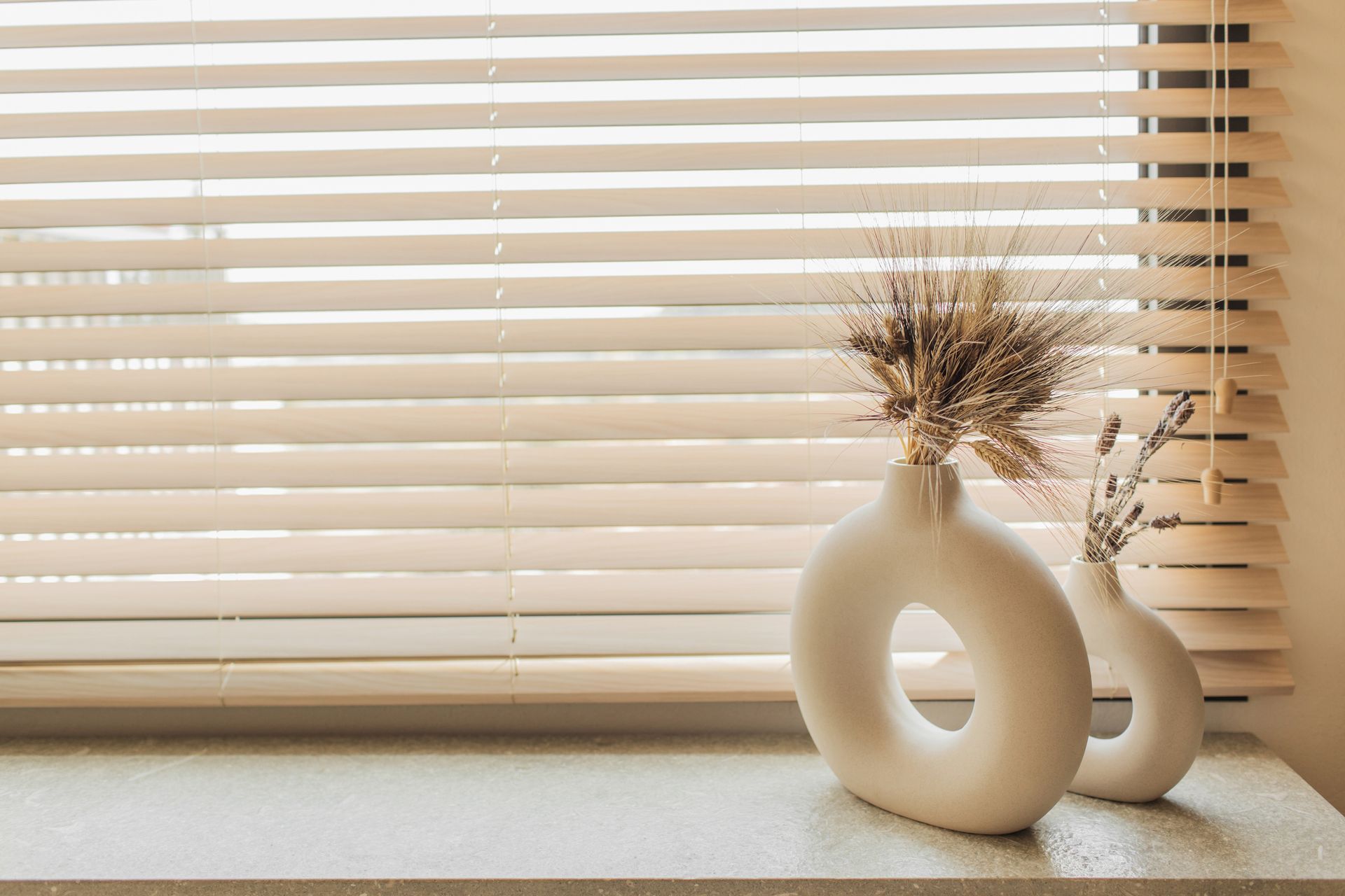 Close-up of decorative vases with dried plants on a windowsill with blinds.