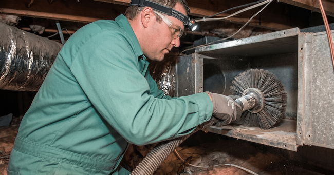 Worker in green coveralls cleaning ductwork with a rotary brush inside a ventilation system.