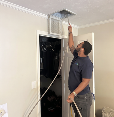 Technician cleaning a ceiling air vent in a hallway, holding a long hose and reaching up with a tool.