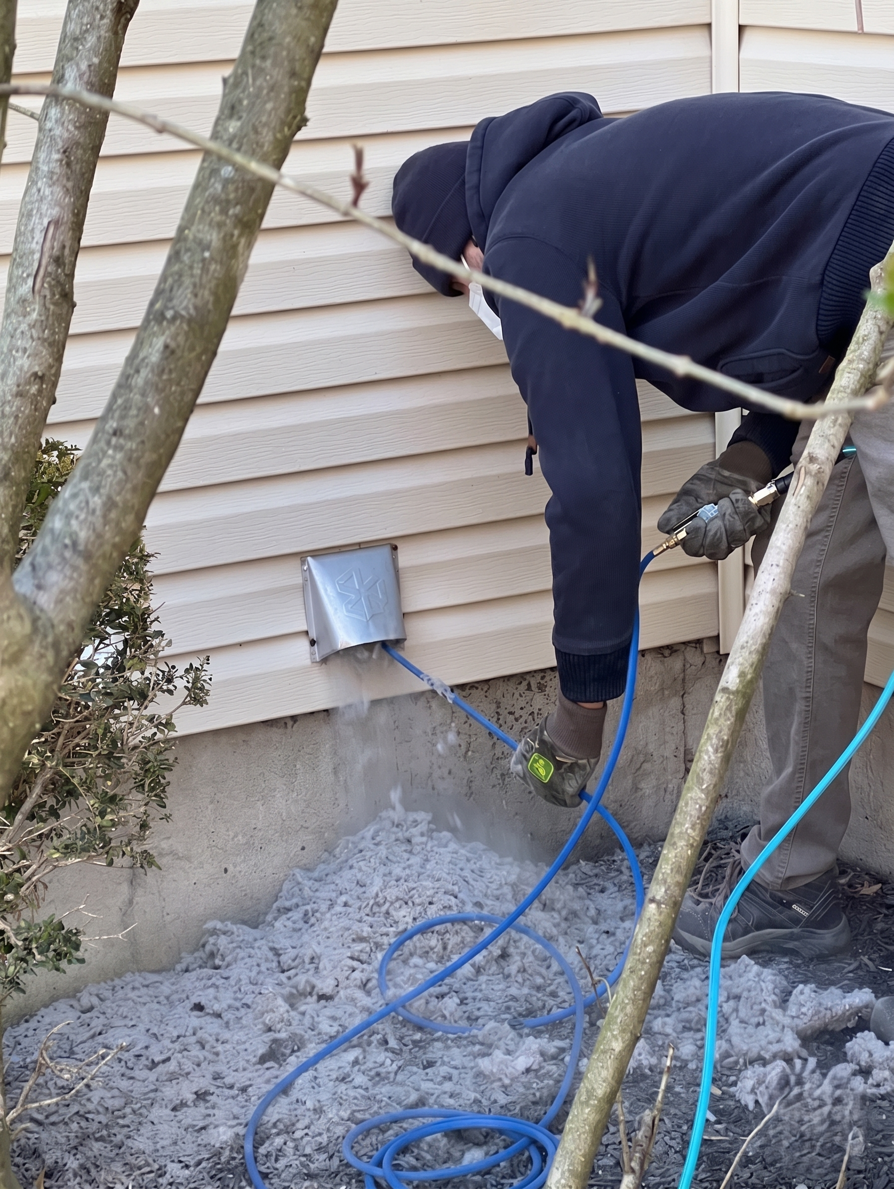 Person in dark clothing using a power tool to work on white house siding near the foundation.