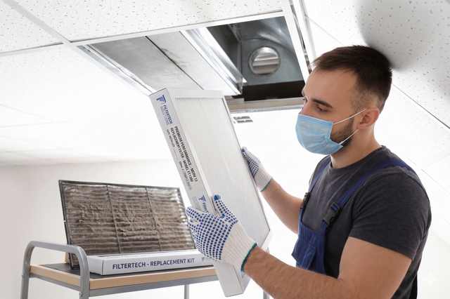 Masked technician replacing an air filter in a ceiling vent indoors