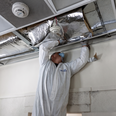 Worker in white coveralls inspecting ceiling ducting and a smoke detector.