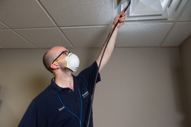 Man wearing a mask and dark scrubs reaching up to inspect a ceiling vent with a flashlight