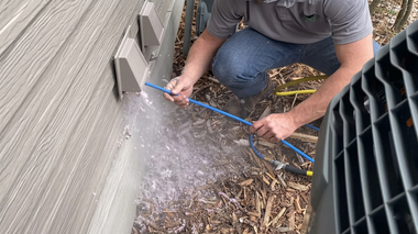 Worker spraying water at the base of a wall with a hose near outdoor HVAC equipment
