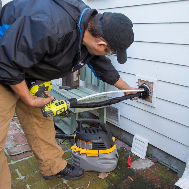 Technician using a drill to install a vent pipe through a house wall beside a vacuum