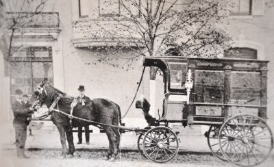 Horse-drawn hearse outside a building. Two men stand near the horse.