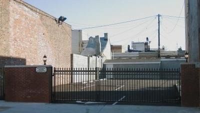 Black gate in front of a brick building. Small sign on left. Overhead power lines and rooftops visible.