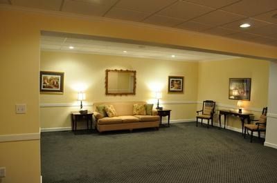 Waiting room with a sofa, chairs, tables, lamps, artwork, and a dark carpet. Yellow walls, neutral setting.