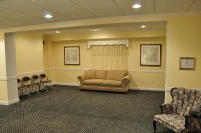Waiting room with beige walls, sofa, chairs, framed art, and carpet.