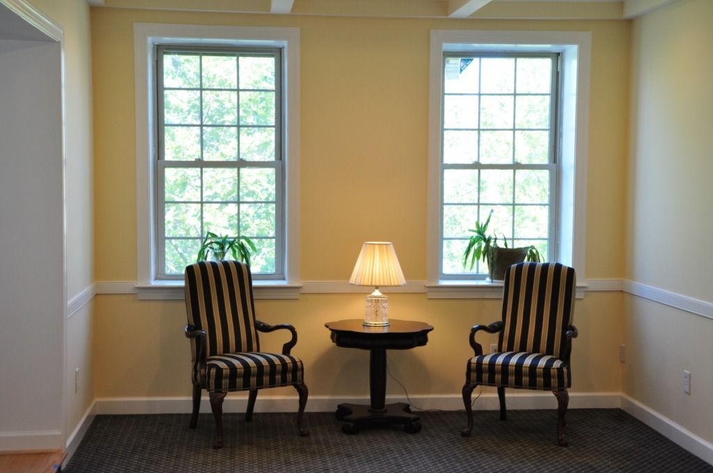 Two striped chairs flank a small table with a lamp, between two windows in a yellow-walled room.