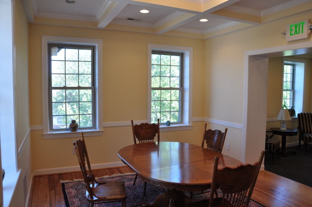 Dining room with oval wooden table, chairs, windows, and exit sign.
