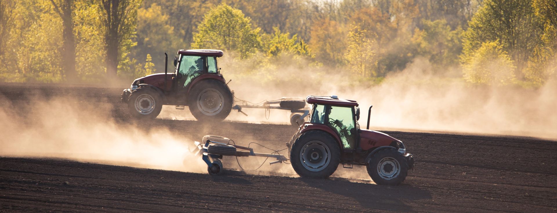Due trattori arano un campo, sollevando polvere; sullo sfondo si vedono degli alberi.