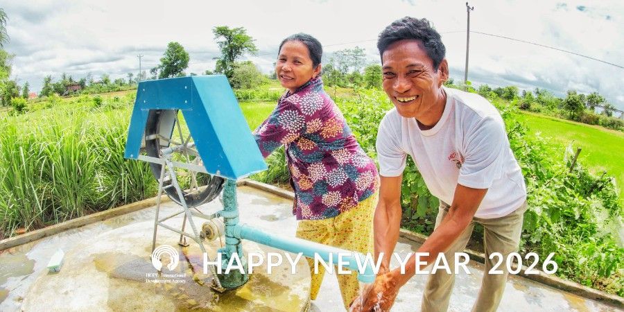Two people near a blue water pump. Smiling man washes his hands. Woman smiles beside the pump. Green fields in the background.