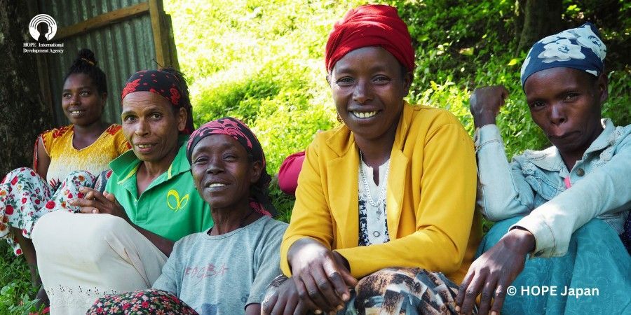 Group of women smiling together outdoors. Some wearing headscarves and bright clothing.