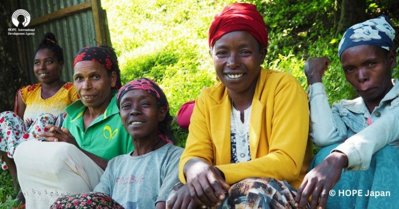 Group of women smiling outdoors, some wearing headscarves, in a sunny environment.